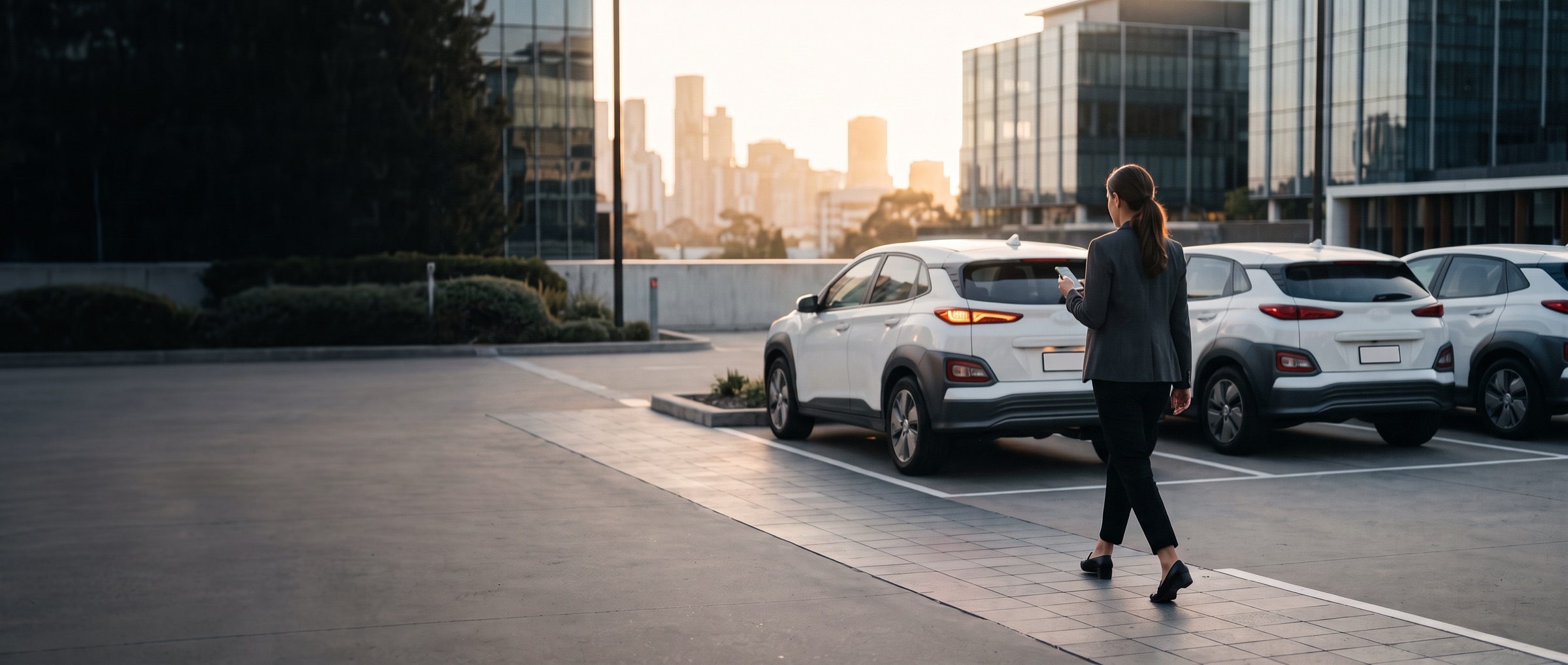 A staff member walks across a city office car park at first light towards a white shared SUV, phone in hand, as its lights come on.